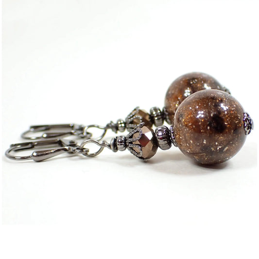 Side view of the handmade galaxy earrings with vintage lucite beads. The metal is gunmetal gray in color. There is a faceted glass bead in metallic brown at the top. The bottom lucite bead is round ball shaped and has marbled shades of brown and black with flecks of glitter and sparkle. Each bottom bead is different from the other for a unique appearance.