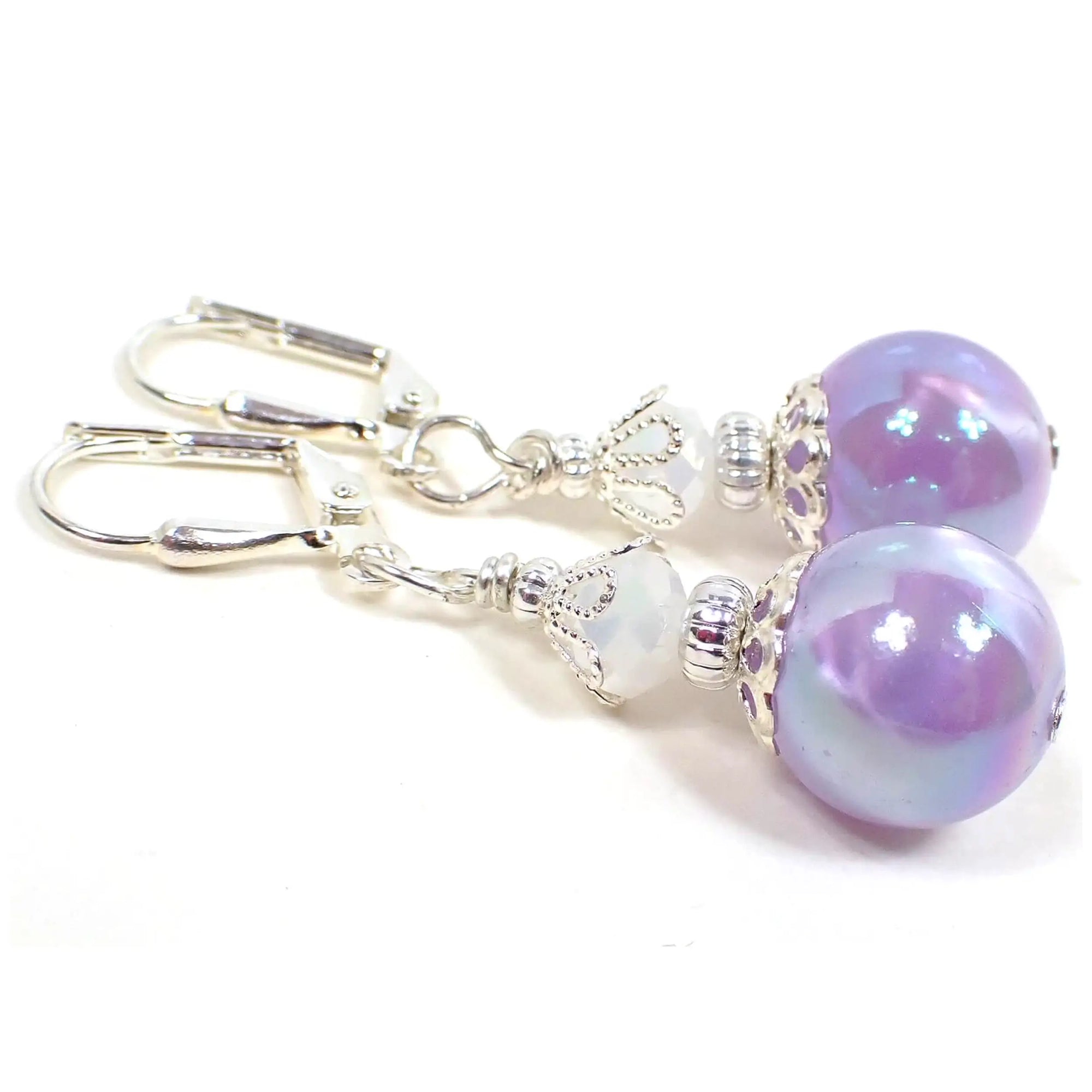 Angled side view of the handmade earrings with iridescent acrylic beads. The metal is silver plated in color. There are faceted white glass beads at the top and pearly iridescent purple acrylic beads at the bottom.