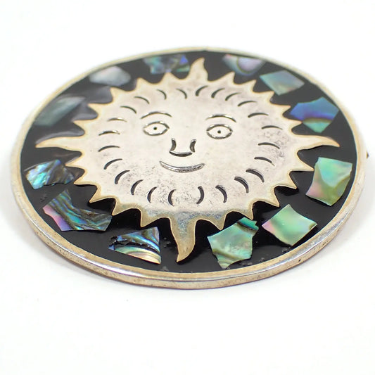 Enlarged front view of the retro vintage Mexican pendant brooch. It's round and has silver tone color metal. There is a sun design in the middle with pieces of inlaid abalone shell around it. The abalone shell has different colors that flash as you move around in the light.