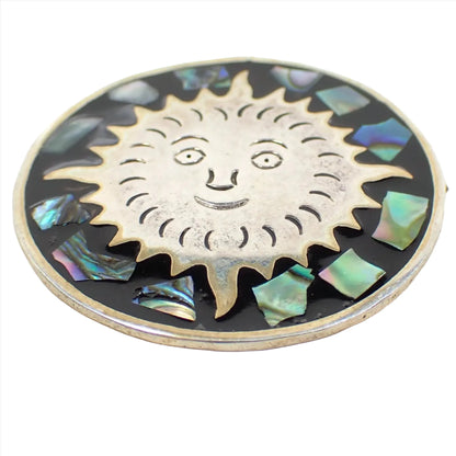 Enlarged front view of the retro vintage Mexican pendant brooch. It's round and has silver tone color metal. There is a sun design in the middle with pieces of inlaid abalone shell around it. The abalone shell has different colors that flash as you move around in the light.