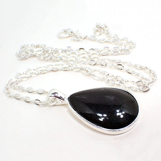 Angled view of the handmade resin teardrop pendant necklace. The chain and setting are silver tone plated in color. The teardrop pendant has a domed pearly black resin cab.