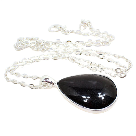 Angled view of the handmade resin teardrop pendant necklace. The chain and setting are silver tone plated in color. The teardrop pendant has a domed pearly black resin cab.