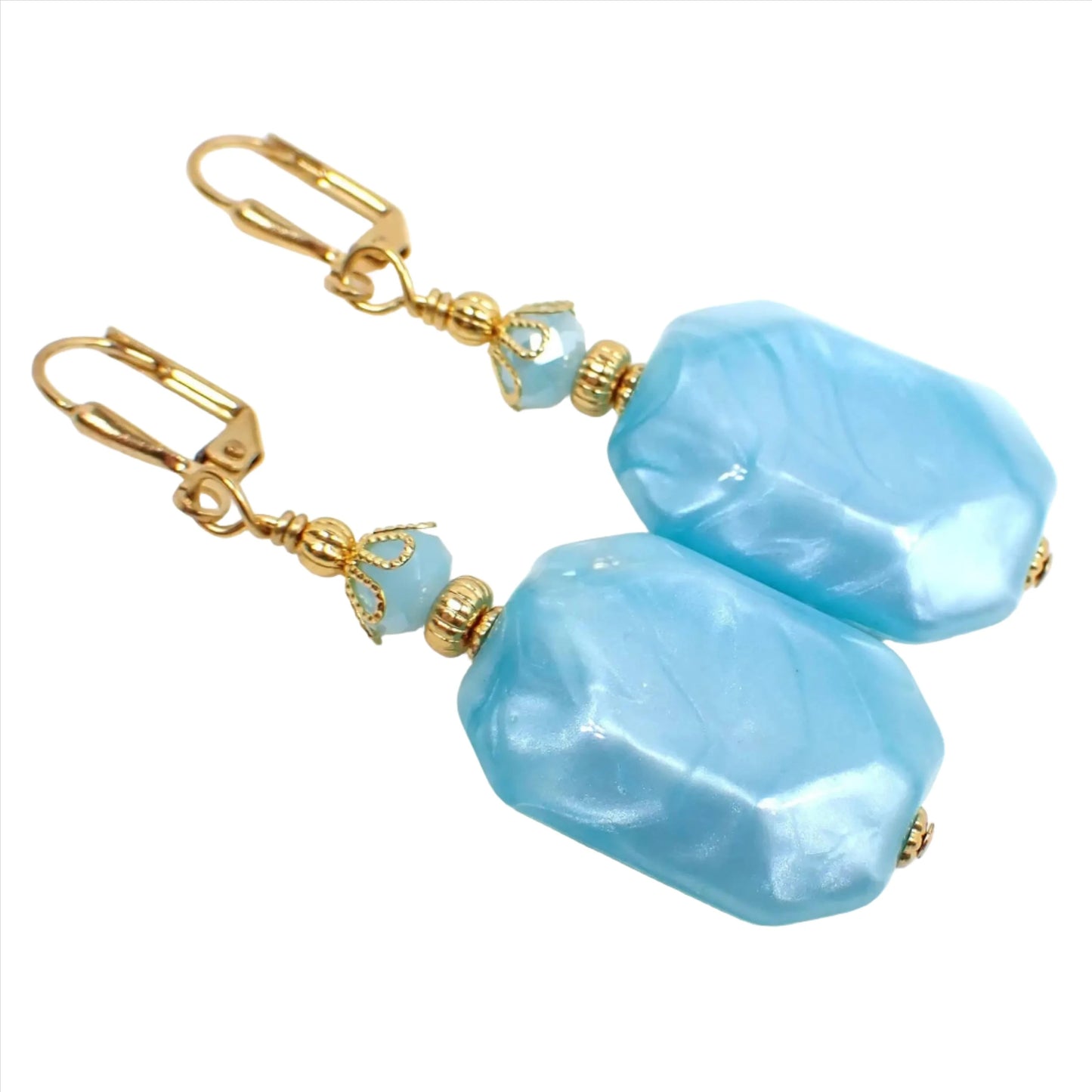 Angled view of the large handmade geometric earrings. The metal is gold plated in color. There are light blue faceted glass beads at the top. The bottom beads are vintage lucite beads that are faceted large octagon shape with a pearly baby blue color.