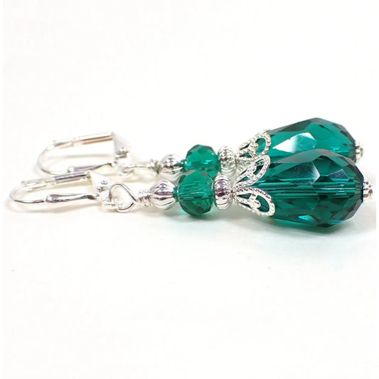Side view of the handmade teardrop earrings. The metal is silver plated in color. There are teal green faceted glass crystal rondelle beads on the top and teardrop shaped beads on the bottom.