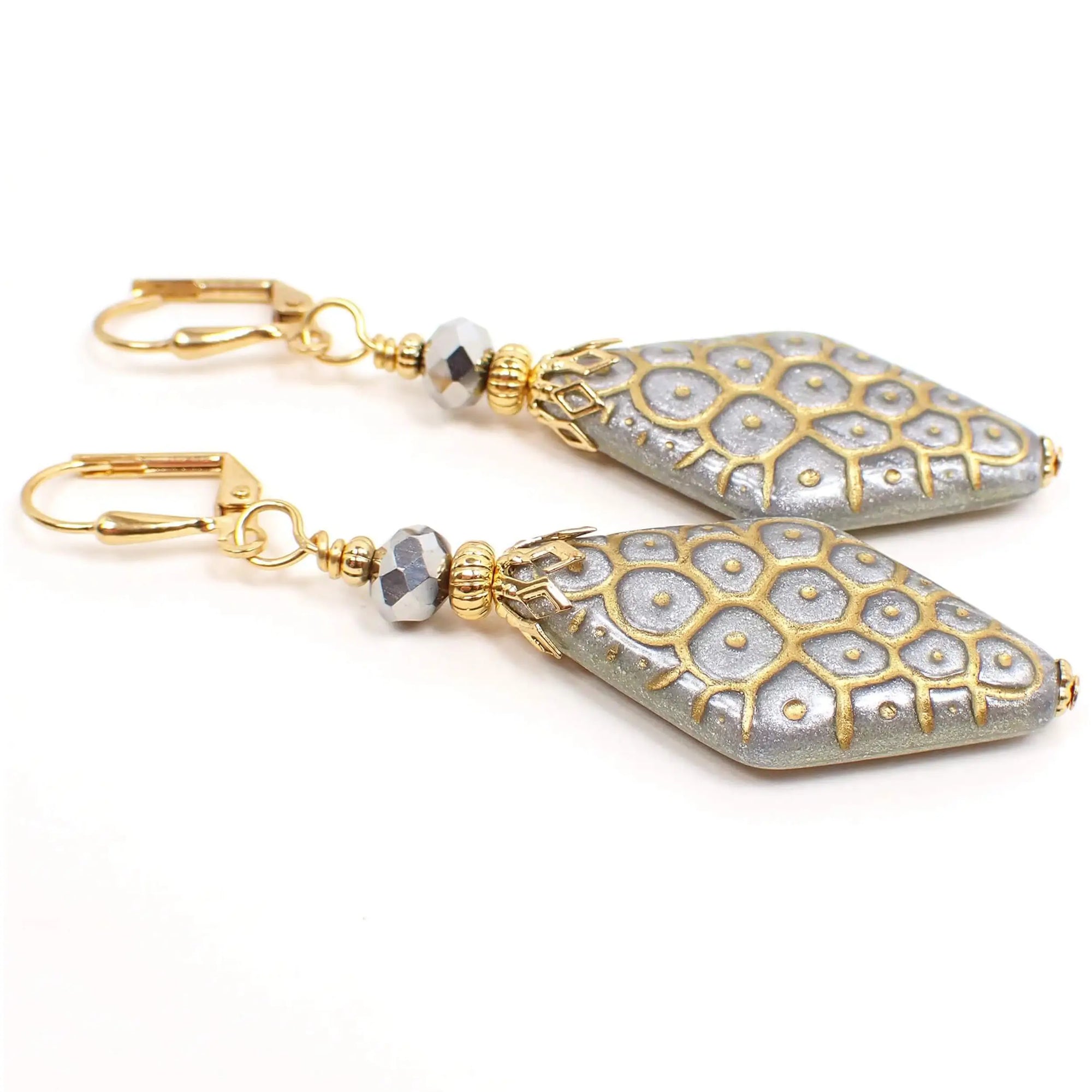 Angled view of the handmade metallic drop earrings made with vintage lucite beads. The metal is gold tone plated in color. There are faceted silver color glass beads at the top. The bottom lucite beads have a puffy flat diamond shape and are metallic silver in color. There is a scale like textured pattern design on them that is in metallic gold color.