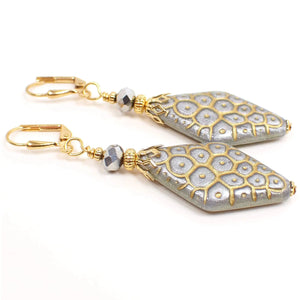Angled view of the handmade metallic drop earrings made with vintage lucite beads. The metal is gold tone plated in color. There are faceted silver color glass beads at the top. The bottom lucite beads have a puffy flat diamond shape and are metallic silver in color. There is a scale like textured pattern design on them that is in metallic gold color.