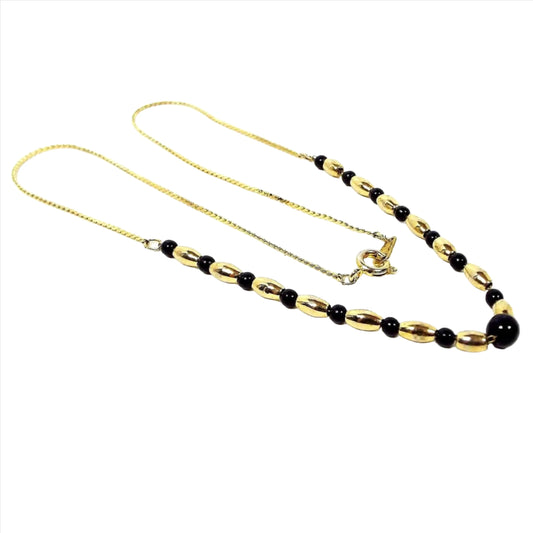 Angled view of the retro vintage beaded chain necklace from South Korea. The metal is gold tone plated. There are gold tone oval metal beads with round black plastic beads on the bottom part of the necklace. The top chain has a serpentine link and spring ring clasp.