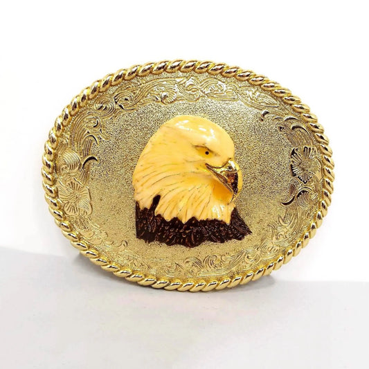 Front view of the retro vintage enameled eagle head belt buckle. The metal is gold tone in color with a floral design around the edge of the oval buckle. In the middle is an enameled bald eagle head with gold tone beak.