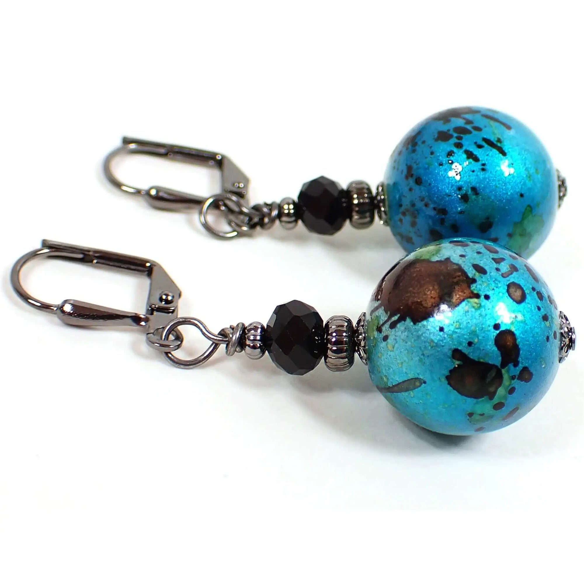 Side view of the handmade drop earrings. The metal is gunmetal gray in color. There are black faceted glass beads at the top. The bottom beads are acrylic and are round ball shaped in a bright metallic teal blue color with black and green splash pattern. Each bottom bead is different from the other in pattern for a unique look.