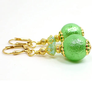 Side view of the handmade sphere drop earrings. The metal is gold plated in color. There is a green faceted glass crystal at the top. The bottom acrylic bead is round with a bumpy texture and is pearly green in color.