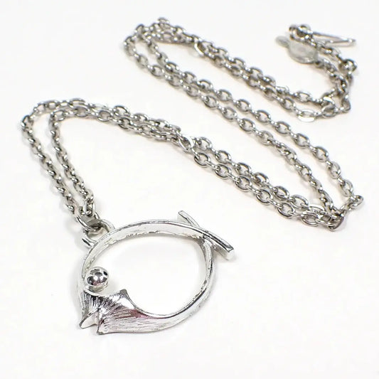 Angled view of the 1976 retro vintage Sarah Coventry "Splash" fish pendant. The metal is silver in color. The chain has a cable link design with a hook clasp and a hang tag seen on the end. The pendant has an open shape like a fish. Some wear to the plating can be seen by the ring at the top and inside area on the pendant under magnification.