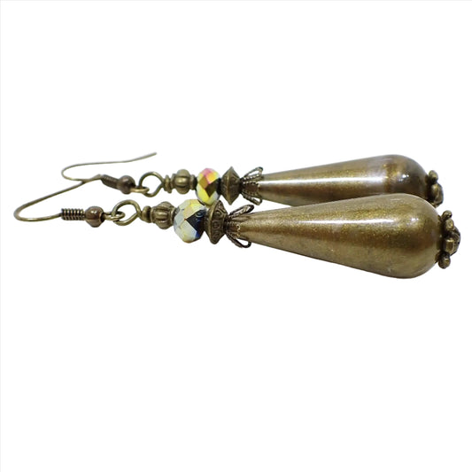 Side view of the handmade teardrop earrings. The metal is antiqued brass in color. There are faceted metallic multi color glass crystal beads at the top. The bottom beads are long teardrop shape and are mostly pearly metallic olive green in color with some hints of brown and other colors swirled in. Each bead is different in color pattern for a unique look.
