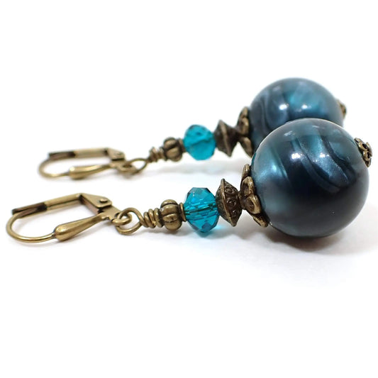 Side view of the handmade teal blue drop earrings. The metal is antiqued brass in color. The top beads are faceted glass crystal in teal blue. the bottom beads are round vintage lucite plastic beads with pearly dark teal blue color.
