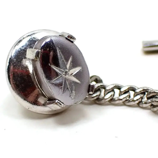 Angled front view of the Swank tie tack. It is oval in shape. Half of the front is matte silver in color and the other half is shiny silver in color. Both silver colors are a darker type silver color. There is an atomic starburst design etched in the middle.