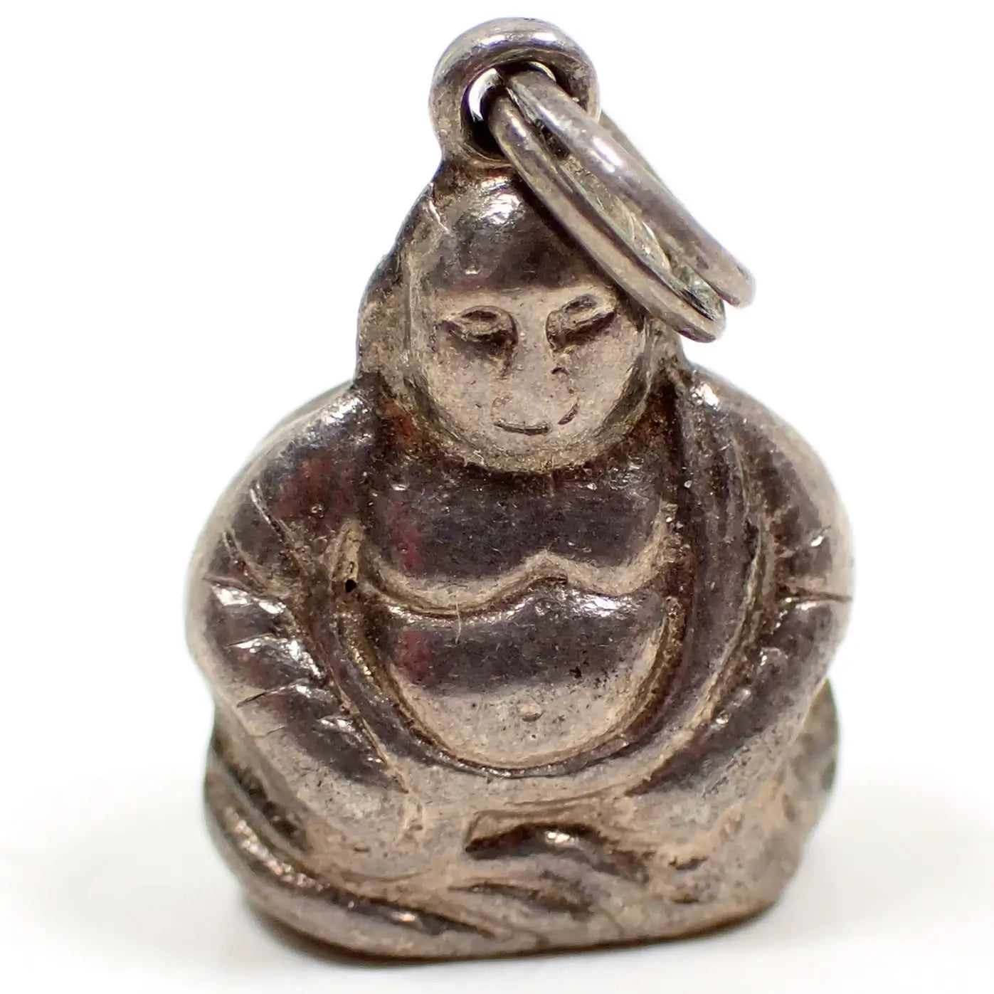 Enlarged front view of the small retro vintage Buddha charm. It is antiqued silver tone in color. There is a loop at the top with a small split jump ring attached to it. The Buddha's face has some rub wear for loss of detail over the years.