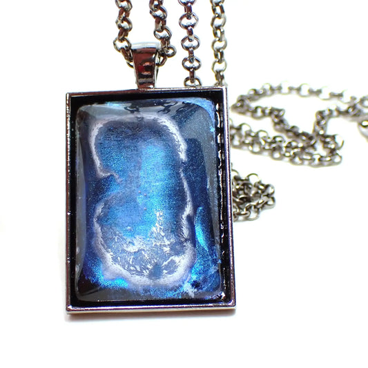 Enlarged front view of the handmade large rectange pendant necklace. The metal is gunmetal plated in color. The resin cab on the front has an iridescent blue background with a white frost like abstract design on top of it.