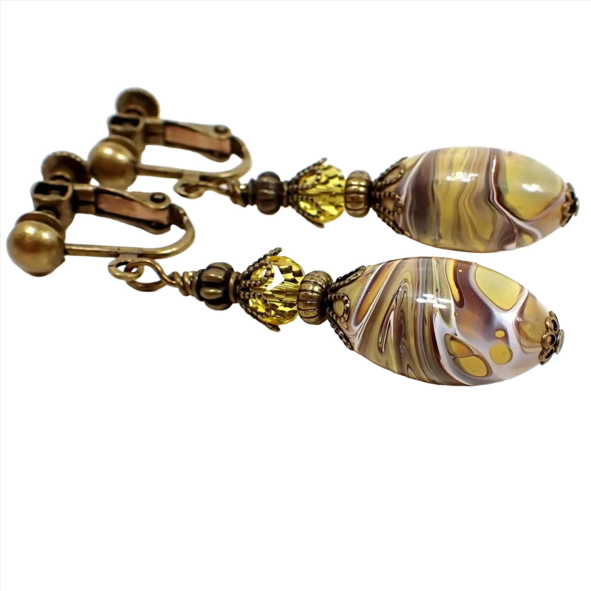 Angled side view of the handmade lucite drop earrings. The metal is antiqued brass in color. There is a faceted yellow glass crystal bead at the top and an oval lucite bead at the bottom that has swirls of white, brown, and mustard yellow.