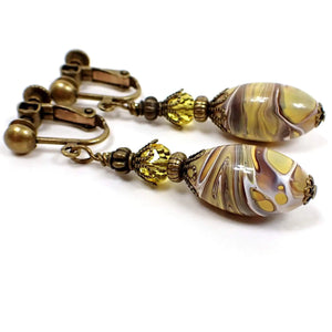 Angled side view of the handmade lucite drop earrings. The metal is antiqued brass in color. There is a faceted yellow glass crystal bead at the top and an oval lucite bead at the bottom that has swirls of white, brown, and mustard yellow.