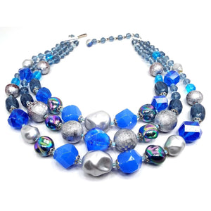 Front view of the Mid Century vintage German multi strand beaded necklace. There are three strands of beads that are made of plastic and are metallic silver and varying shades of blue. There are round, oval, and nugget shaped beads that go from smallest to largest as you go down the necklace.