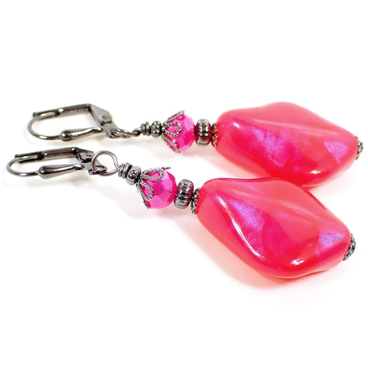 Angled view of the handmade earrings with vintage color shift lucite beads. The metal is a dark gray gunmetal. There are hot pink faceted glass beads at the top. The bottom beads are lucite, are shaped like wavy diamond shapes with rounded edges. They are a bright pink in color with pearly hints of purple as you move around in the light.