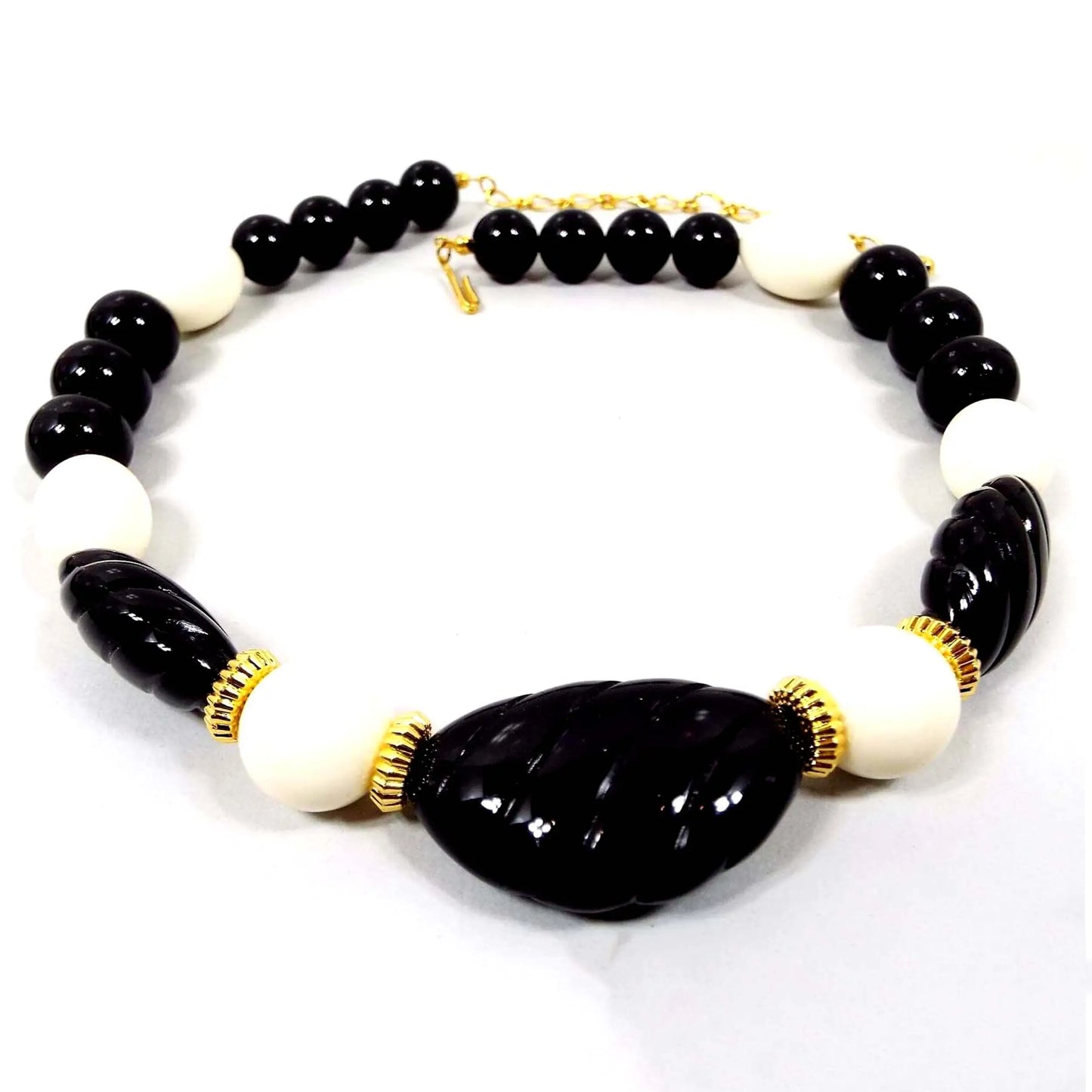 Front view of the retro vintage black and white lucite beaded necklace. The metal is gold tone in color. There are various sizes of round, and oval shaped lucite plastic beads in black and white. There is a chain at one end and a hook on the other to adjust the length. The metal is gold tone in color.