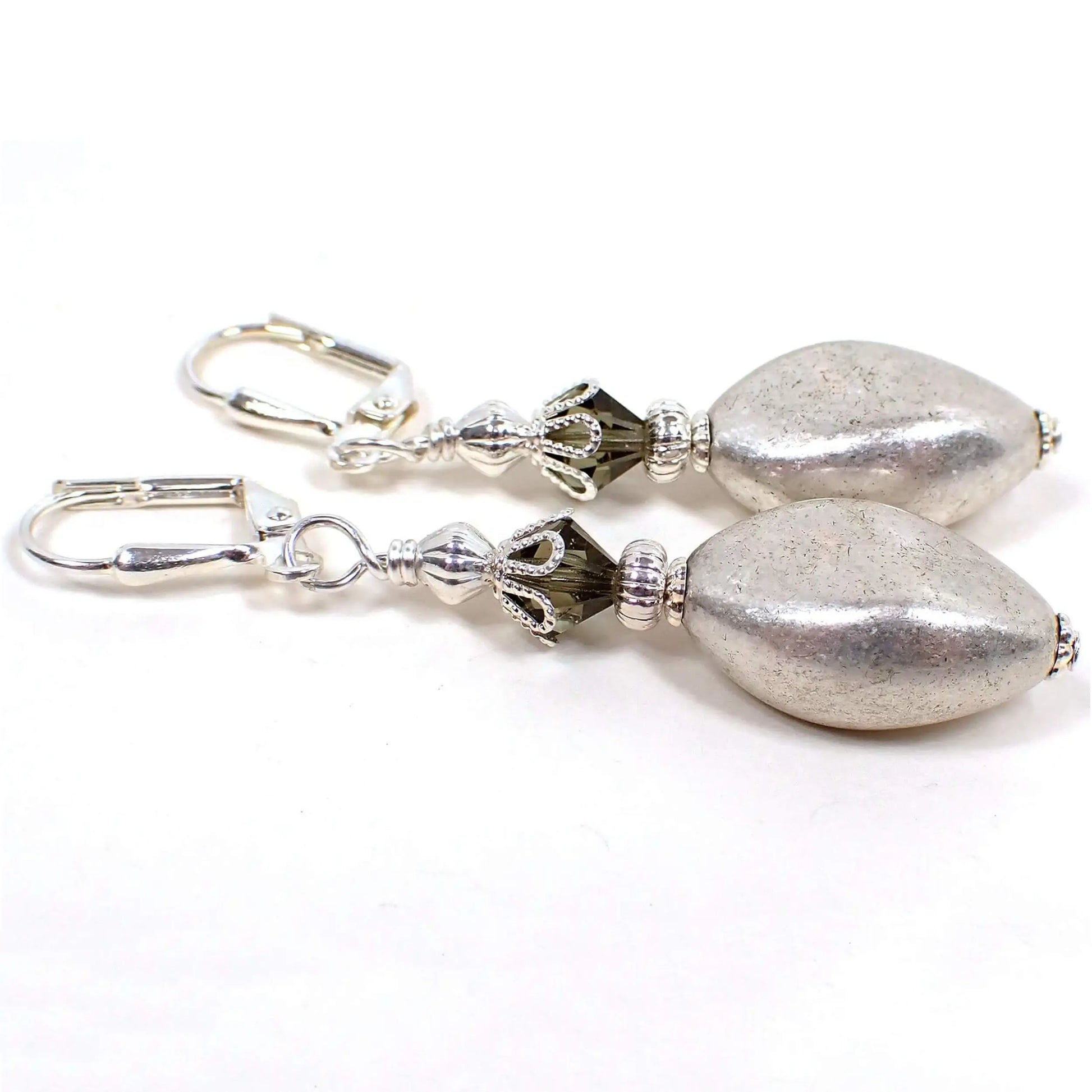 Side view of the handmade drop earrings. The metal is silver plated in color. There are brownish gray faceted glass crystal beads at the top. The bottom acrylic beads are a twisted oval shape and are metallic silver in color.