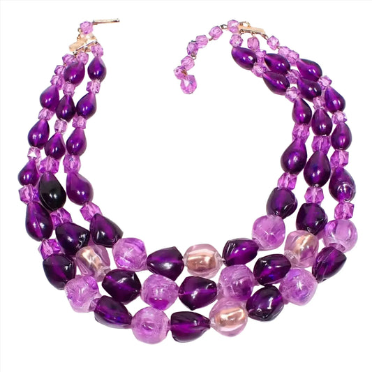 Top view of the Mid Century vintage Western Germany multi strand necklace. There are three beaded strands with various chunky plastic beads in shades of purple. There is a hook clasp and chain at the end to make it slightly adjustable in length.