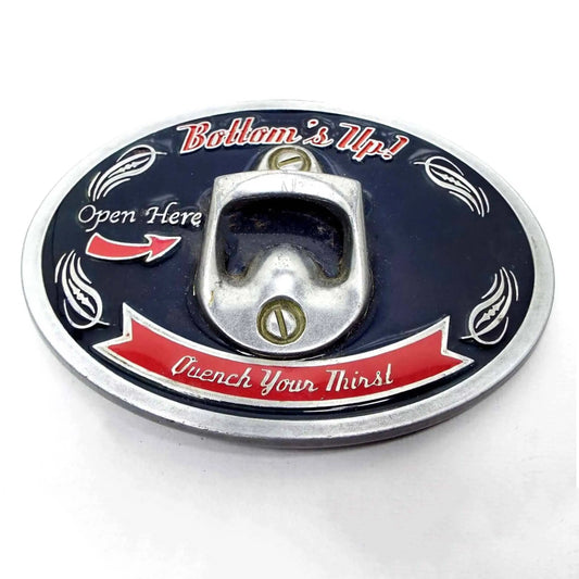 Front view of the retro vintage novelty belt buckle. It's oval and silver tone in color with dark blue enamel background and red enameled areas where the lettering is. There is a bottle opener in the middle. It says Bottom's Up, Open Here, and Quench Your Thirst on it.