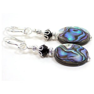 Angled view of the handmade abalone earrings. The metal is silver tone plated in color. There is a black faceted glass crystal bead at the top. The bottom abalone shell beads are flat oval shaped and have shades of gray, purple, blue, and green that have some flash as you move around in the light.