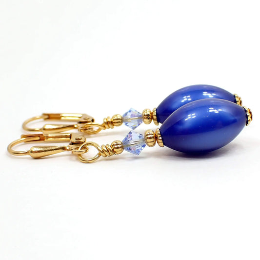 Side view of the small handmade lucite drop earrings. The metal is gold plated in color. There is a light blue faceted glass crystal bead at the top. The bottom bead is a smaller sized oval moonglow lucite bead in deep blue. The moonglow lucite has a glowy effect on the inside of the bead as you move around in the light.