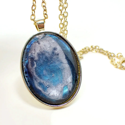 Enlarged front view of the handmade large oval pendant necklace. The metal is gold plated in color. The resin cab on the front has an iridescent blue background with a white frost like abstract design on top of it.