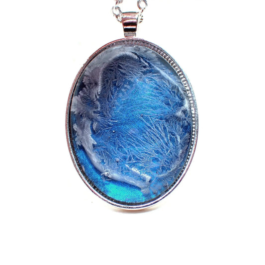 Enlarged front view of the handmade large oval pendant necklace. The metal is silver plated in color. The resin cab on the front has an iridescent blue background with a white frost like abstract design on top of it.