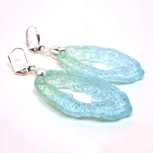 Angled front view of the handmade faux geode slice earrings. The metal is silver plated, They are long oval shaped with an open middle. The edges have a druzy like appearance. The resin is semi translucent blue with some green at the top. There are tiny specks of blue glitter throughout.