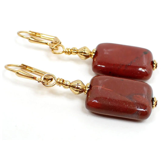 Angled view of the handmade red jasper earrings. The metal is gold plated in color. There are two metal beads at the top. The bottom jasper bead is puffy rectangle shaped with rounded corners. It's primarily a dark brick red in color with a few areas of other colors marbled in.