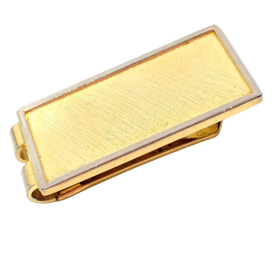 Angled top view of the retro vintage money clip. It is shaped like a rectangle and has a brushed matte gold tone color front with a silver tone color edge.
