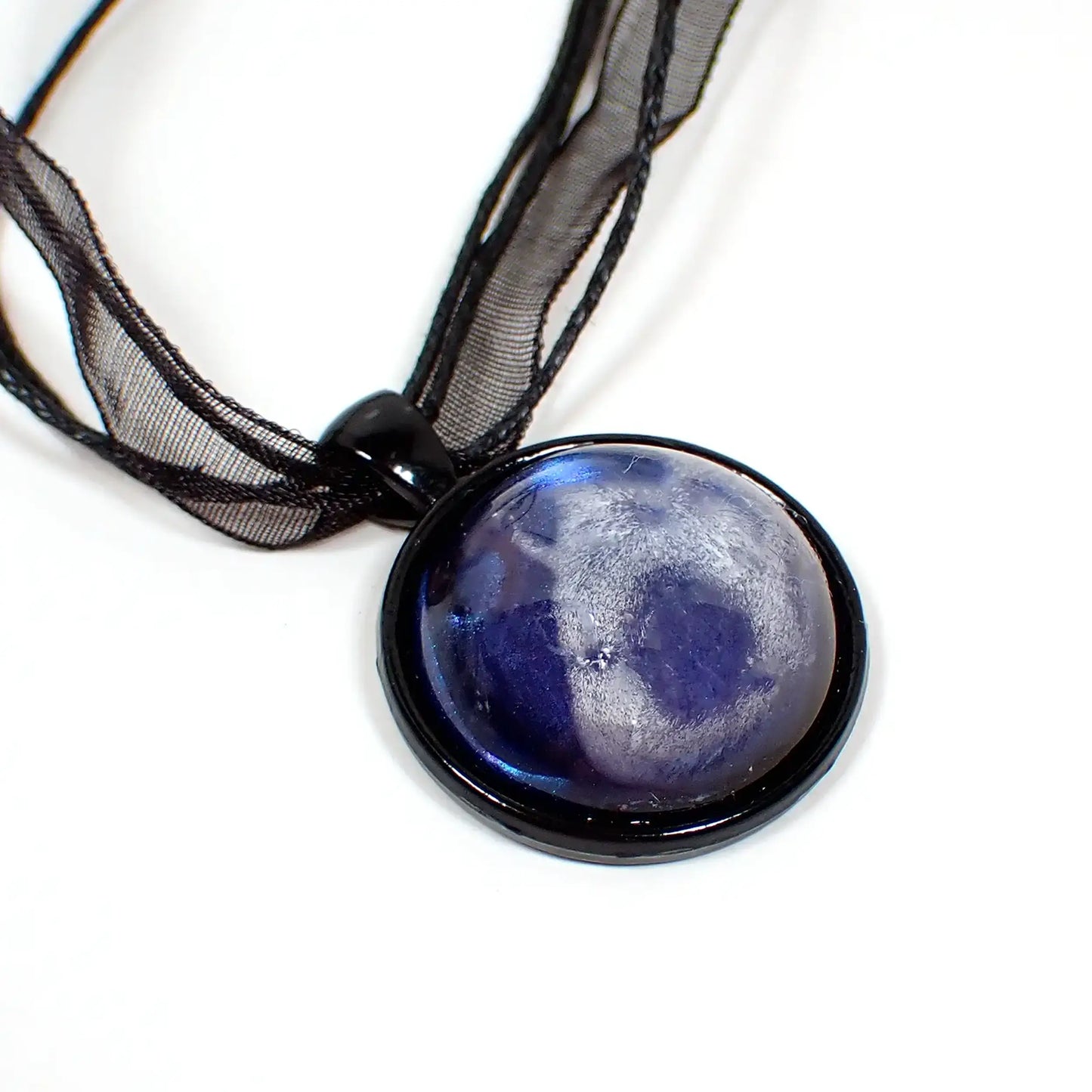 Enlarged view of the handmade black and resin pendant necklace. The necklace has three black strands of faux leather cord and a strand of organza ribbon. The pendant has a black coated base and is round. There is a domed round cab on the front made of resin with an iridescent blue background and a white abstract frost like design going through it.