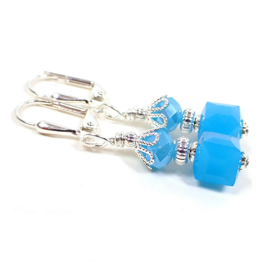 Angled view of the handmade cube earrings. The metal is silver plated in color. There are bright mint blue faceted glass rondelle beads at the top and square cube beads at the bottom.