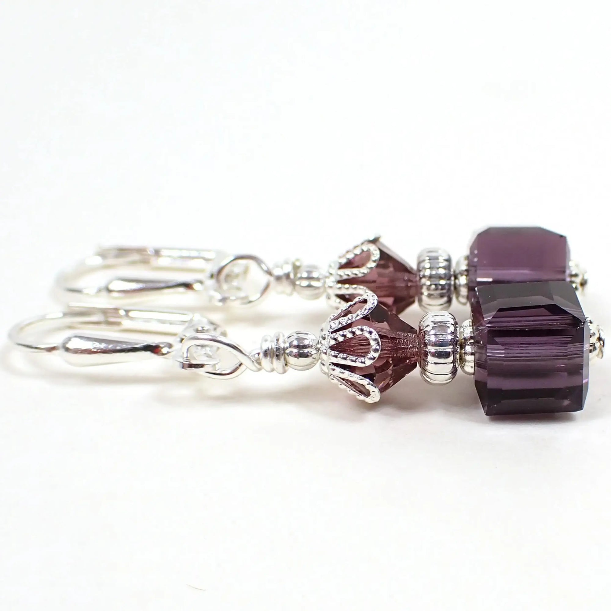 Side view of the handmade cube drop earrings. The metal is silver plated in color. There are faceted glass crystal bicone beads at the top and square cube beads at the bottom. The beads are dark purple in color.