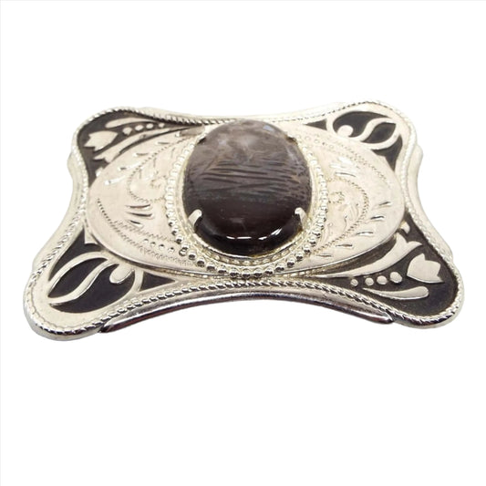 Top view of the retro vintage agate gemstone belt buckle. It is silver tone metal with a black painted floral design on the corners. In the middle is an oval agate gemstone cab that has shades of dark gray and brown. The area around the cab h as an etched leaf like design.