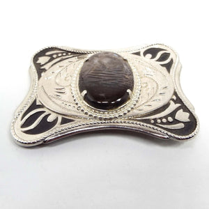 Top view of the retro vintage agate gemstone belt buckle. It is silver tone metal with a black painted floral design on the corners. In the middle is an oval agate gemstone cab that has shades of dark gray and brown. The area around the cab h as an etched leaf like design.