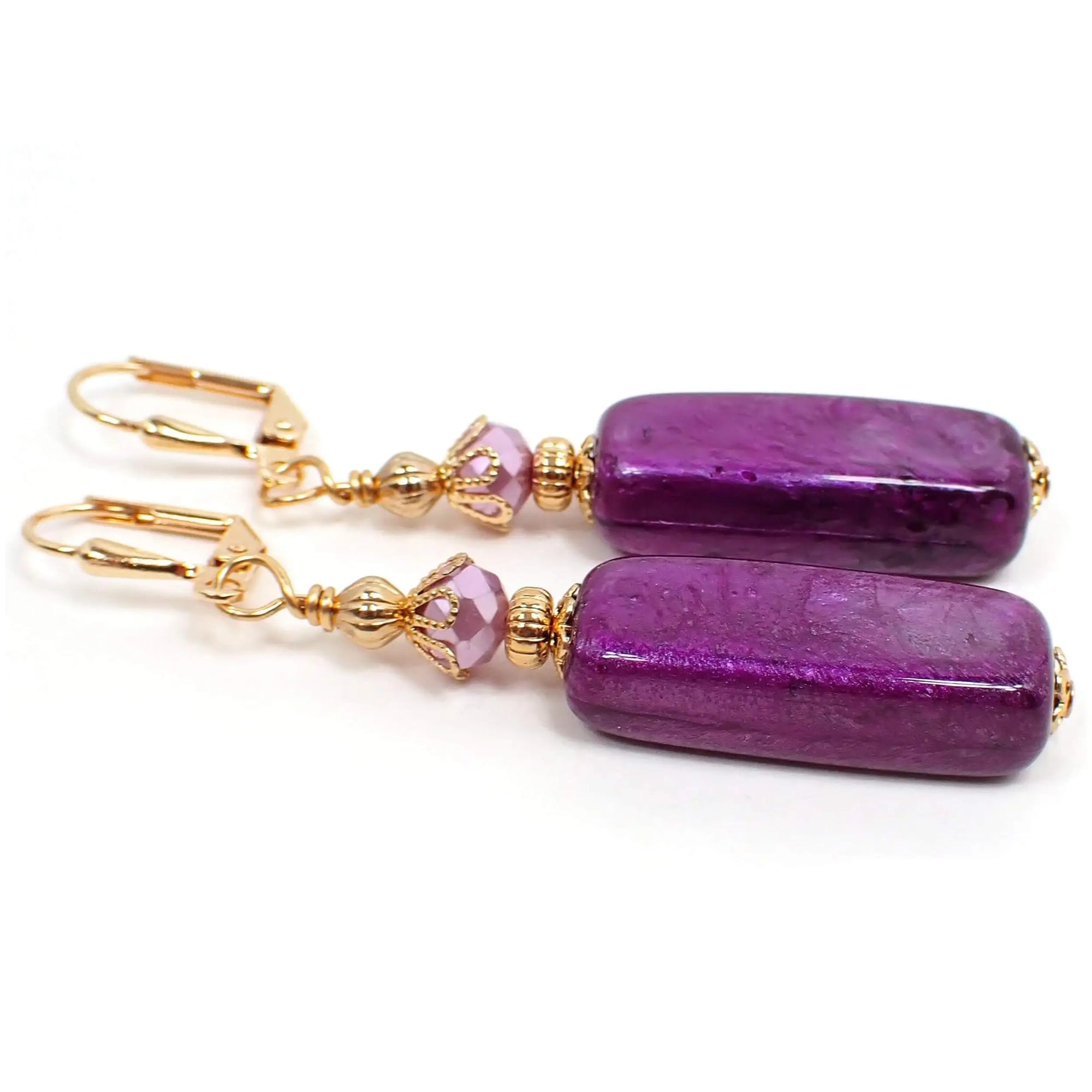 Side view of the handmade large stick earrings made with vintage acrylic beads. The metal is gold plated in color. There are faceted glass crystals at the top of the earrings in an opaque pastel purple color. The bottom acrylic beads are long larger sized acrylic beads with a pearly vivid purple color.