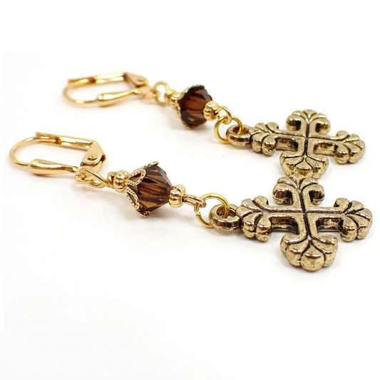 Angled view of the handmade cross earrings. The metal is gold plated in color. There are brown faceted glass crystal beads at the top. The bottom cross charms are antiqued gold plated and have an even cross design with flared out scalloped ends.