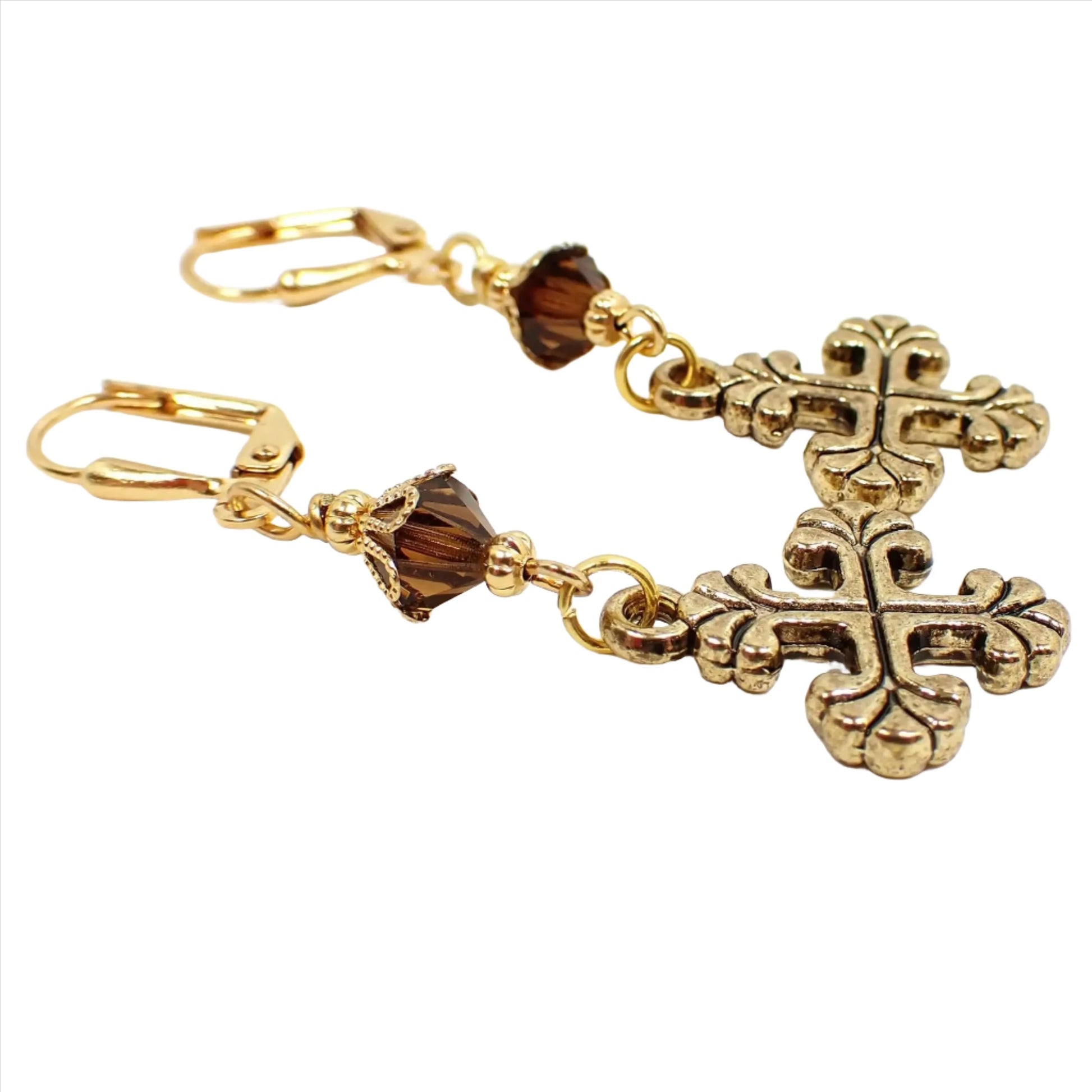Angled view of the handmade cross earrings. The metal is gold plated in color. There are brown faceted glass crystal beads at the top. The bottom cross charms are antiqued gold plated and have an even cross design with flared out scalloped ends.