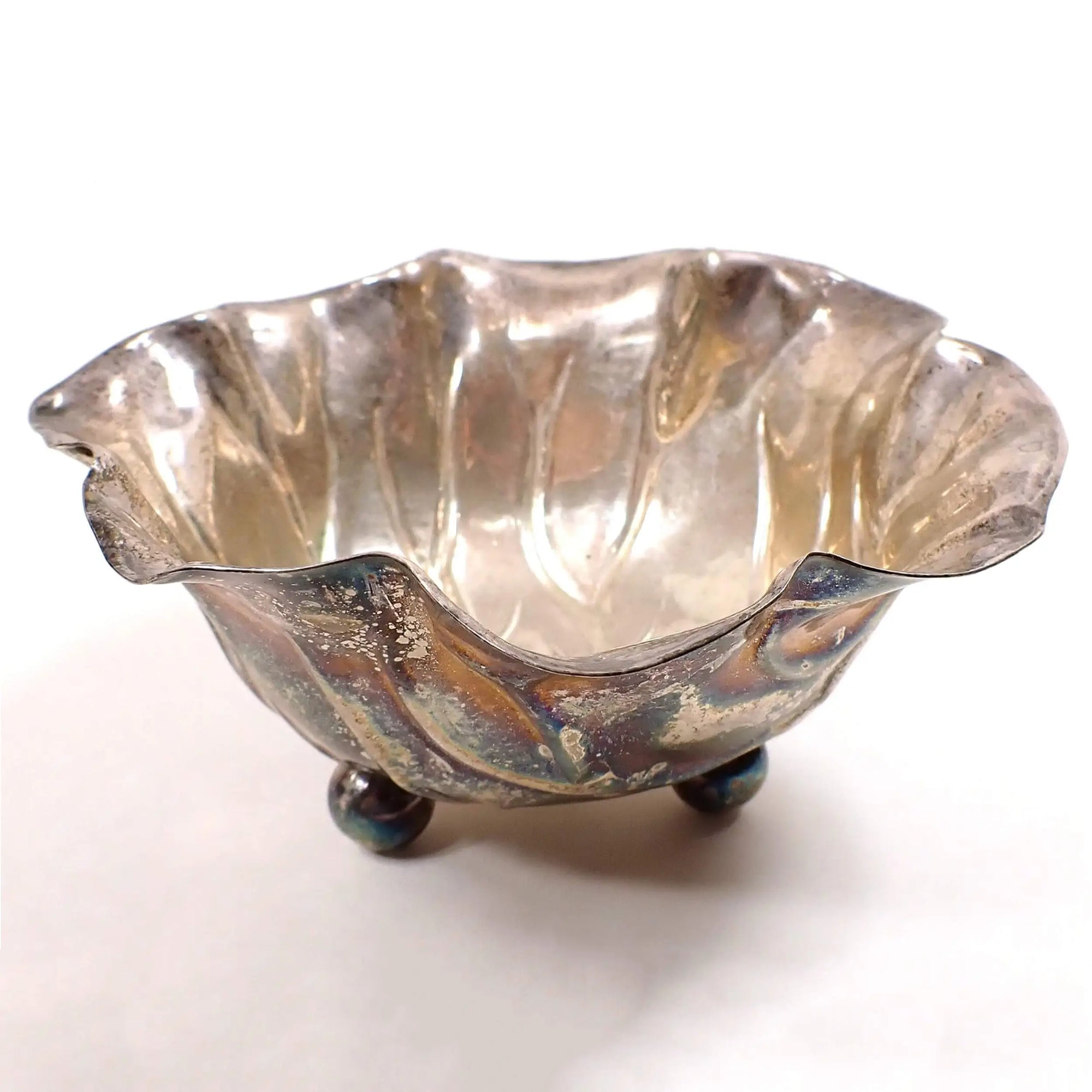 Angled view of the retro vintage Mexican Alpaca bowl. It is completely made out of metal and is a darkened antiqued silver tone in color. The bowl part is shaped like a cabbage leaf and there are three round ball "feet" at the bottom of the bowl.