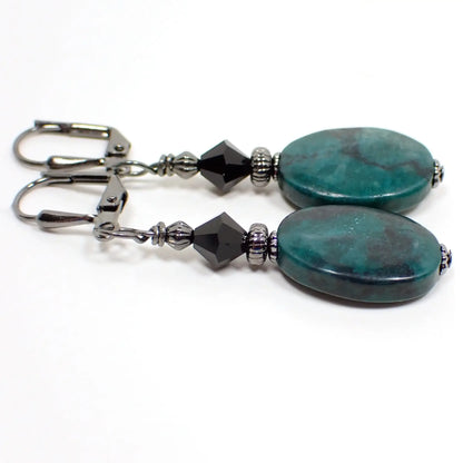 Side view of the handmade gemstone earrings. The metal is gunmetal gray in color. There are black faceted glass crystal beads at the top. The bottom beads are flat oval shaped and are primarily teal green with black marbled in.