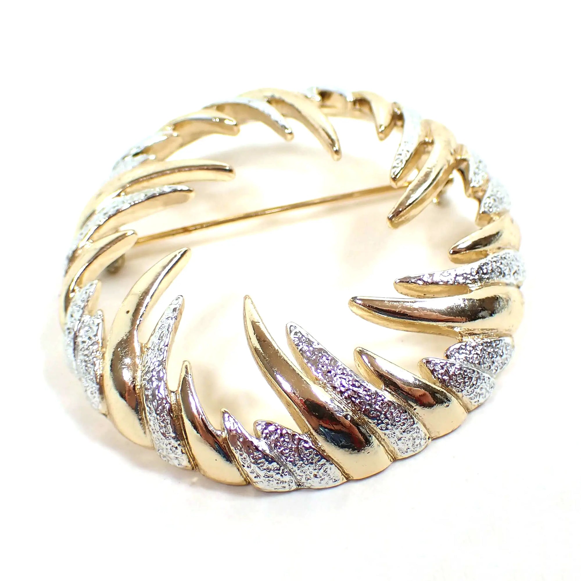 Front view of the retro vintage Sarah Coventry brooch pin. It is round in shape with an open middle. The is a curved spike like design going inward all the way around the brooch. The colors rotate between shiny gold tone plated and textured matte silver tone plated color.
