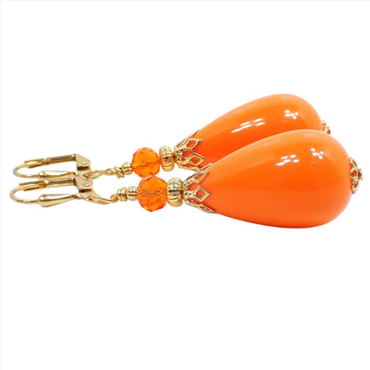 Side view of the large chunky handmade teardrop earrings. The metal is gold plated in color. There is a bright orange faceted glass crystal bead at the top of the earrings. The bottom bead is a vintage lucite bead that is teardrop shaped and also bright orange in color.