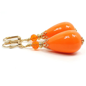 Side view of the large chunky handmade teardrop earrings. The metal is gold plated in color. There is a bright orange faceted glass crystal bead at the top of the earrings. The bottom bead is a vintage lucite bead that is teardrop shaped and also bright orange in color.