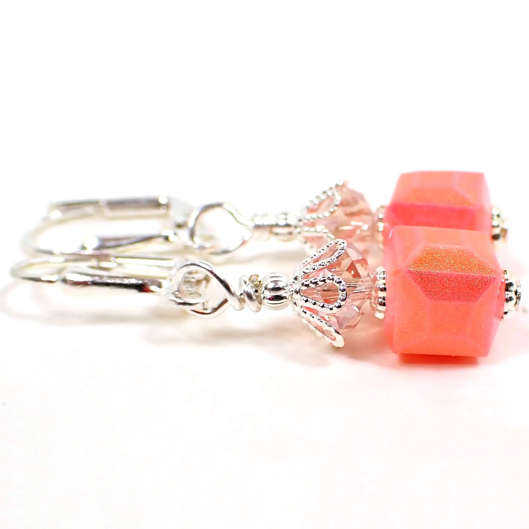 Side view of the small handmade cube earrings. The metal is silver plated in color. There are light pink faceted glass crystal beads at the top. The bottom beads are square cube shaped and have a bright pink color with a sparkly golden sheen on the outside.