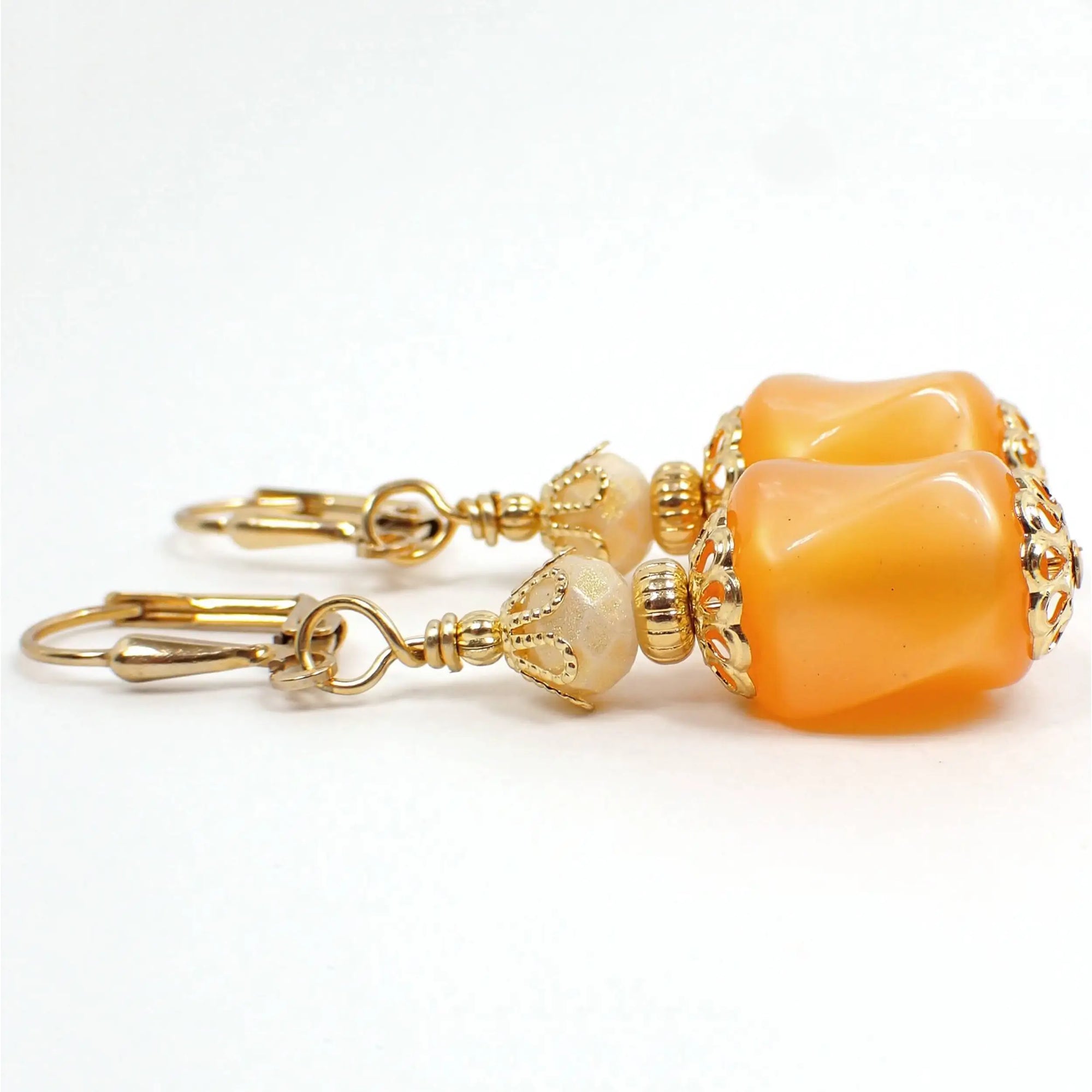 Side view of the handmade drop earrings. The metal is gold plated in color. There are new faceted glass crystal beads at the top that are off white with a light golden sheen on them. The bottom beads are vintage moonglow lucite beads in peach color. They are twisted barrel shaped and have an inner glowy like appearance as you move around in the light.